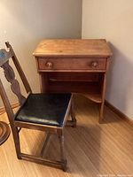 View of the vintage small wooden desk with one drawer and lower shelf, showing surface and drawer knobs, alongside the vintage chair with black padded seat.