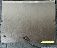 Front view of the black textured art portfolio with artist name label on bottom right corner, showing the exterior with tie closure.