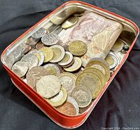 Tin container filled with assorted international coins and banknotes, viewed from above showing various coins and folded bills.