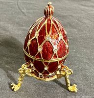 Front angled view of red enamel Faberge style egg trinket box showing netted pattern with rhinestone accents and gold-tone base with claw feet.