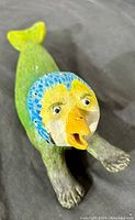 Close-up photo of the bird/fish hybrid ceramic figurine showing the blue and yellow bird-like head, with a yellow beak and green fish tail, resting on a black surface.