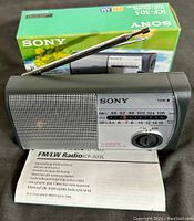 Sony ICF-303 portable radio with extended antenna, original box behind, instruction manual below. Front face shows speaker grill and tuning dial with frequency display panel.