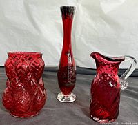 Three cranberry glass vases displayed side by side on a gray surface with a white backdrop. The items show the textured vase, tall etched vase with a clear base, and cranberry glass pitcher with clear handle.