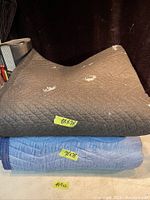 Two folded moving blankets stacked, dark gray on top, blue underneath with visible size labels.