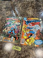 Photo showing two vintage Superman comic books on a textured surface. The comic on the left is DC Comics Presents issue #57 featuring Superman and Robin. The comic on the right is Superman issue #369. Both show signs of normal wear with visible creases and some edge wear.