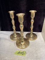 Photo of three solid brass candle holders arranged on a white surface with a black background. Each candle holder is about 7 1/4 inches tall with a rounded base and classic turned stem design.