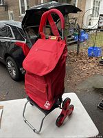 Red folding trolley dolly showing front view with padded handles, zippered flap bag, and distinctive tri-wheel assembly on a driveway setting.