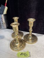 Photo of three solid brass candle holders placed on a surface with a dark background. The candle holders have round bases and tapered tops for holding candles.
