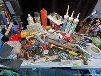 Wide view showing large assortment of hand tools, hardware, and spray cans on table.