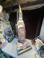 Silver upright Bissell vacuum cleaner standing on a grey table, showing front with clear dust container and brand labeling.