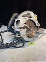 Side view of silver and black Mastercraft circular saw showing blade and handle with power cord coiled, with normal wear.