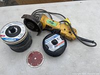 DeWalt angle grinder with attached power cord alongside three different abrasive discs on a white table surface.