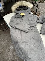 Front and side view of a grey TNA winter coat with a fur-lined hood, lying on a white table outside showing texture, pockets, and zipper details.