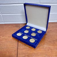 Blue velvet presentation box opened showing eight silver-colored collectible coins each labeled with Alaska-themed subjects.
