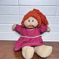 Full body photo of the vintage Cabbage Patch Doll sitting against a white textured background, showing the pink dress and beige cloth limbs.