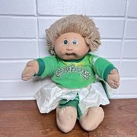 Front view of vintage Cabbage Patch Kids doll sitting against a white brick wall, showing green top with logo and yarn hair.
