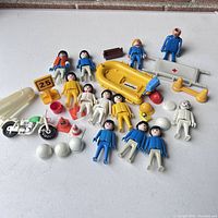 Photo showing vintage PLAYMOBIL figures, a yellow lifeboat, motorcycle, traffic cones, stretcher, helmets and accessories spread on a white background