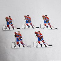 Top view of five tin hockey player cutouts depicting Montreal hockey players in blue, red and white uniforms, holding sticks in skating poses, laid out on fabric.