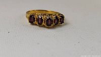 Close-up front view of gold-tone ring with five oval purplish-red garnets on white background.