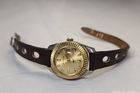 Side angle showing gold-tone dial, 10k solid gold fluted bezel, and brown leather strap with three circular holes on each side.