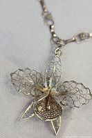 Close-up of silver-tone filigree orchid pendant showing detailed lace-like petals and 3D layered design.