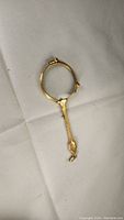 Gold-tone magnifying lorgnette lying on a white surface, showing handle, circular lens, hinge/clip, and size.