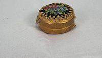 Small round box with pettipoint floral decal on lid with ornate gold-tone metal edging and smooth gold-tone metal body