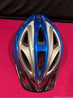 Front view of large blue and silver Alpina bicycle helmet with ventilation and black visor against black and pink background.
