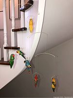 Wide view of bird mobile hanging near staircase, showing five hand-carved and painted birds in vibrant colors suspended on wire frames.