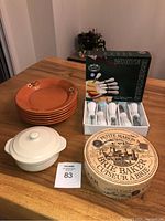 Overall lot showing terracotta plates stacked, white porcelain Brie baker with lid, cheese cutter box, and round brie baker container