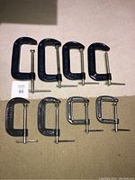 Full view of all 8 steel C-clamps arranged in two rows on a carpeted floor, showing different sizes.