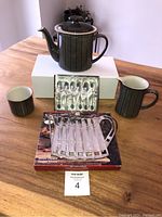 Photo of full set on a wooden table showing teapot, creamer jug, cup, abalone spoons in box, and silver plated trivet in box.