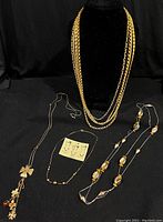 Four gold-toned fashion necklaces displayed on black background and jewelry bust, showing various chain styles and lengths, including heavy multi-strand chain and beaded necklaces.
