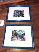 Two colorful framed prints depicting Nova Scotia harbour scenes with boats, houses, and lighthouse. Displayed on wooden surface.