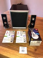 Three items shown: HP speaker system with subwoofer and two satellite speakers on left and right, two Legrand USB chargers on packaging in front, and boxed emergency LED bulb on far right.