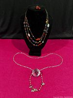 Three-strand colorful glass bead necklace with matching red bead earrings on black bust; long crystal bead necklace with large faceted crystal pendant displayed below on magenta surface.
