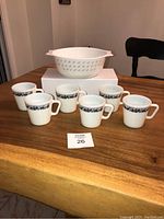 Six white vintage Pyrex mugs with dark blue Old Town Blue Onion pattern around exterior, arranged on table, with white casserole dish in background