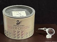 Photo showing the original cylindrical Swarovski Silver Crystal box with swan logo and branding next to the crystal kiwi figurine on a dark surface.