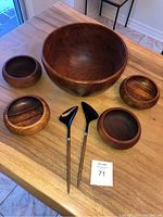 Full view of all items on wooden table: large teak bowl, five smaller teak bowls, and black nylon tipped salad servers with teak handles and stainless steel shafts.