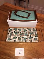 Three vintage serving trays: one mosaic tile tray with metal handles and two turquoise Formica wood trays, placed on a wooden table and a box for display.