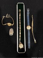 Overview of three women's vintage wristwatches and two watch bands arrayed on a surface, including gold plated, silver-tone, rectangular, round with blue strap, and a chain link watch band