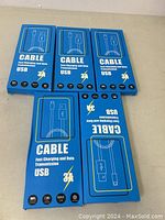 Five sealed blue retail packs labeled 'CABLE Fast Charging and Data Transmission USB 3A' with icons showing 100% satisfaction, fast charging, max 3A, and nylon material. Each pack appears to contain 2 cables.