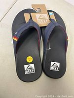 Pair of new men's Reef flip flops in black with dark gray straps and rainbow stripe detail, size 10, manufacturer tags attached.