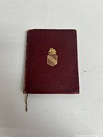 Front view of small red vintage manuscript book with gold emblem on cover, showing textured cover material.