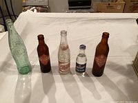 Front view of five vintage soda bottles on white surface including two Orange Crush, Pepsi, Coke, and Schweppes bottles.