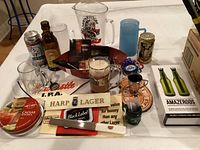 Photo showing assorted bar items on a white table including glass mugs, pitcher, beer brand signs, bottle openers, a metal tray and a blue plastic cup