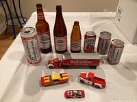 Photo of Budweiser collection showing five Budweiser glass beer bottles, four aluminum cans, and three Budweiser-themed toy vehicles arranged on a white cloth surface.