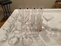 Photo showing 15 assorted cocktail glasses including martini glasses, highball glasses, a measurement glass, and shot glasses arranged on a white tablecloth
