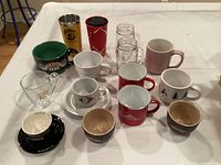 Photo of 15 assorted coffee cups and mugs in various colors and designs on a white tablecloth.