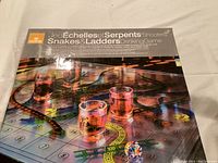 Game box cover showing Snakes and Ladders drinking game with shot glasses and multicolor die on board.